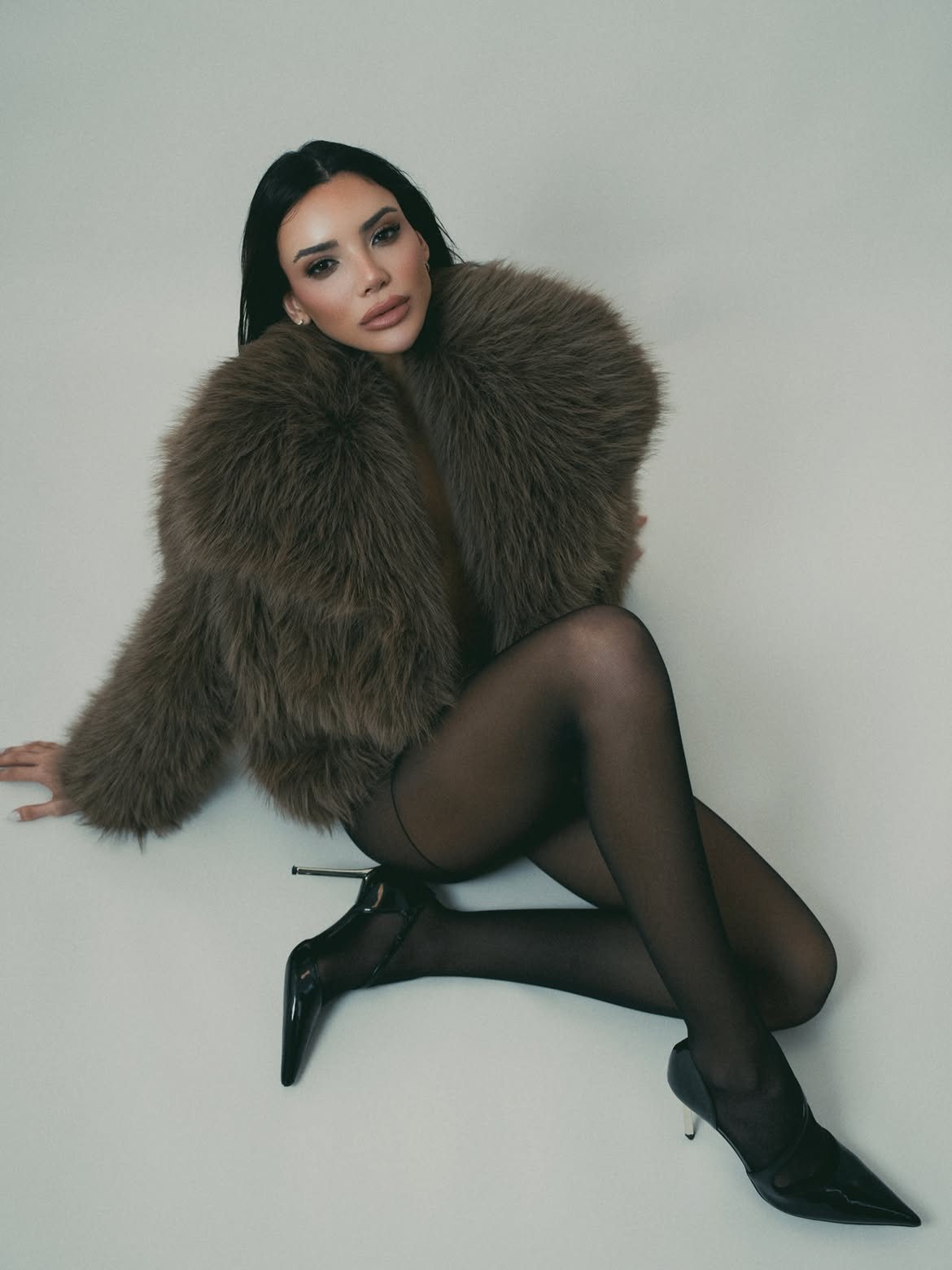 Eileen León seated on the studio floor in a brown fur jacket and sheer black tights