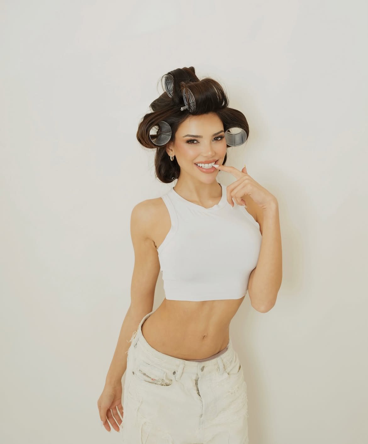 Eileen León in hair rollers and a white crop top, smiling
