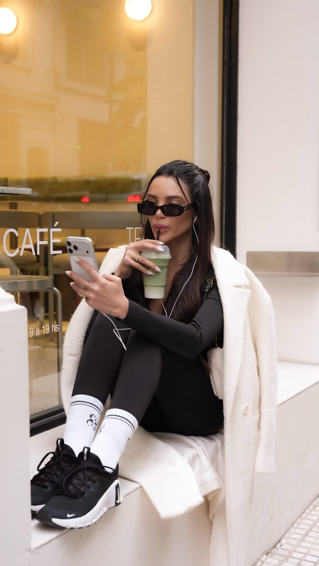 Model in black Eiloa set and white coat sipping a matcha outside a café, phone in hand
