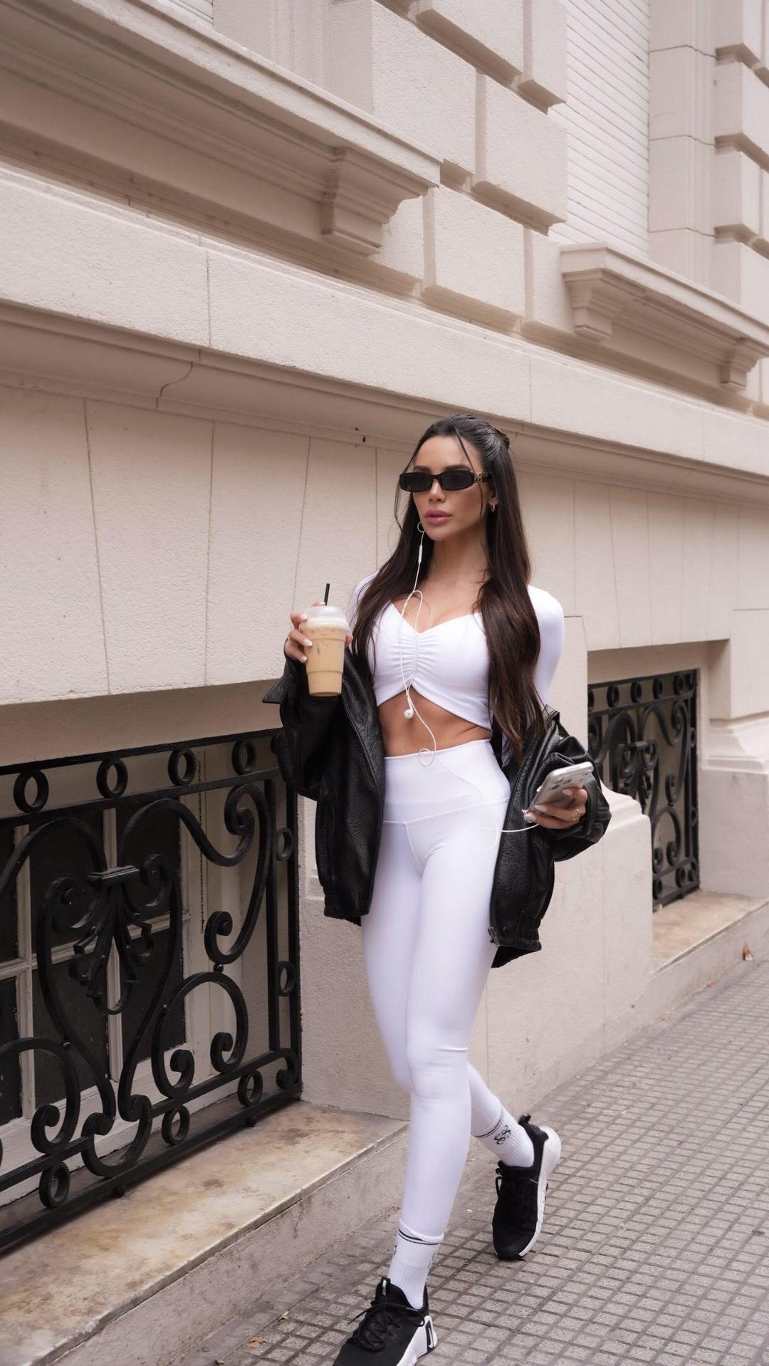Model in white Eiloa set and black leather jacket walking past wrought-iron windows with an iced coffee
