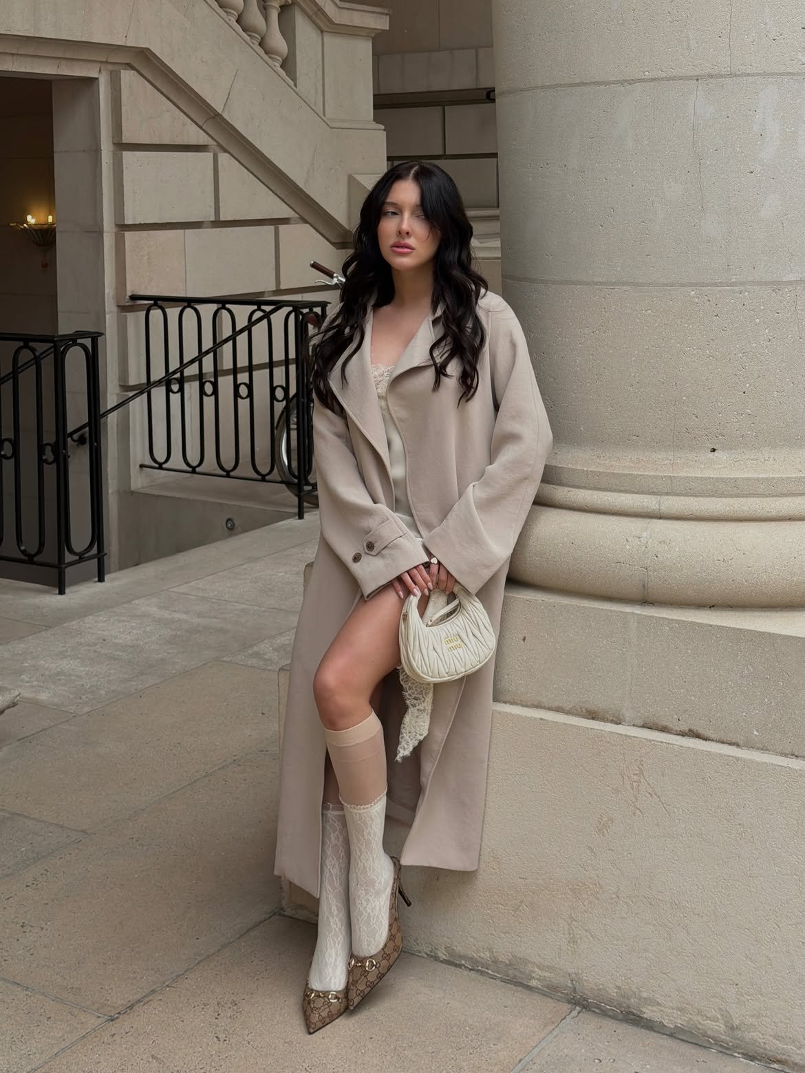 Model in a beige trench and lace socks holding a Miu Miu bag against a stone column