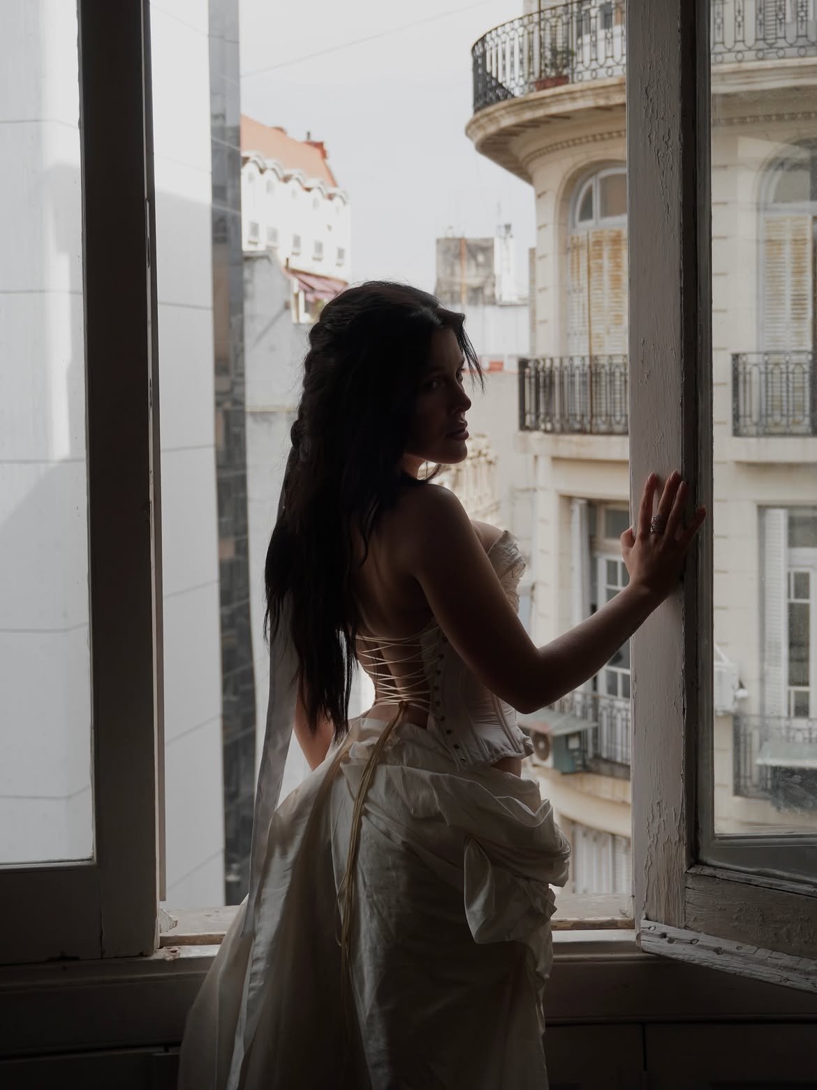 Model in an ivory corset at an open window, back to camera, Buenos Aires balconies beyond