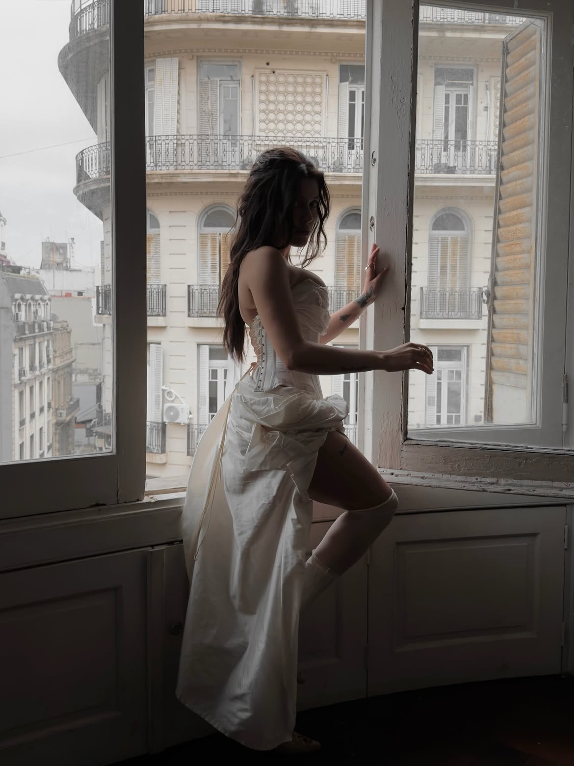 Model in an ivory corset stepping onto a window ledge, silhouetted against a curved façade