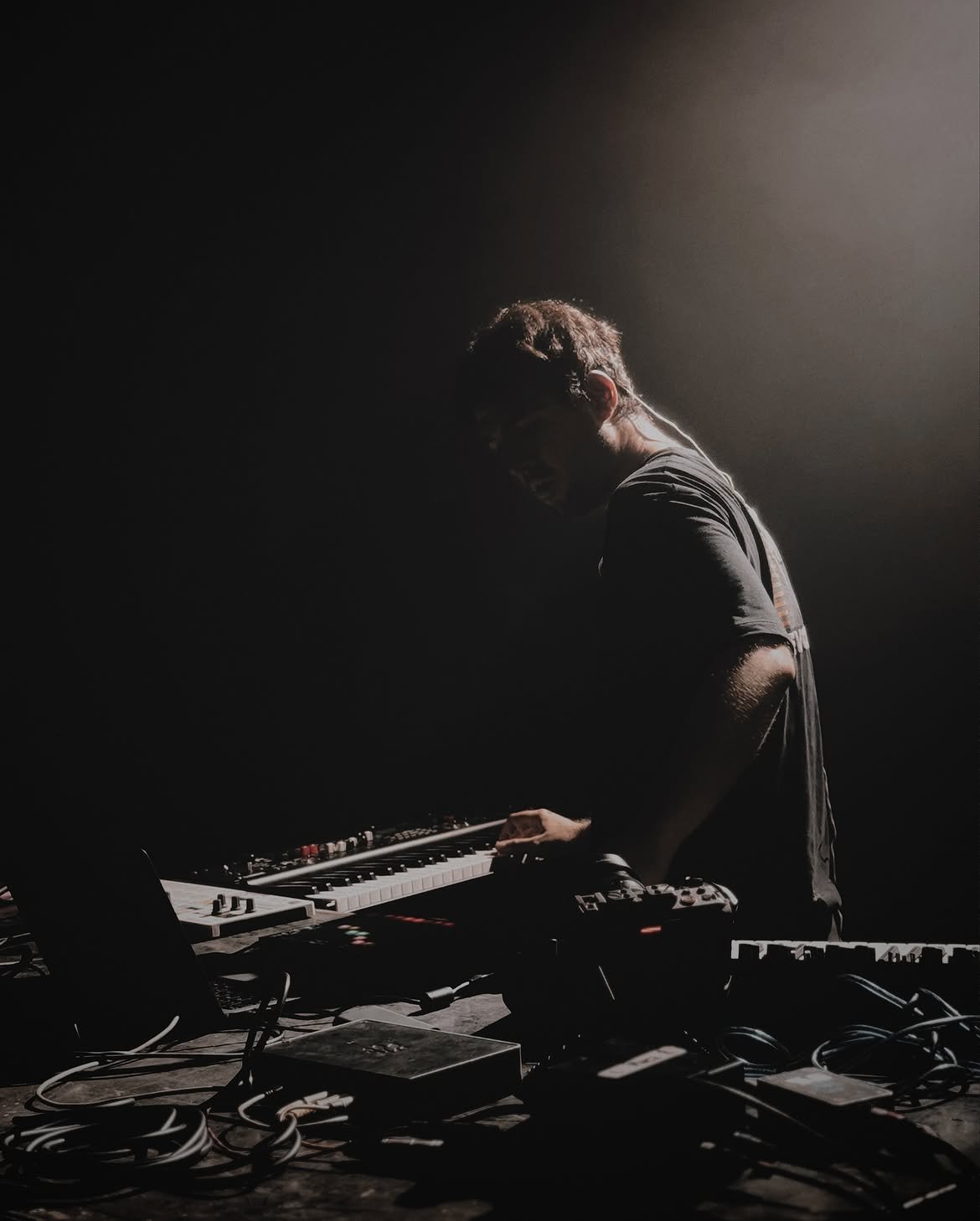 Musician at a synthesizer on stage, backlit by a single spotlight