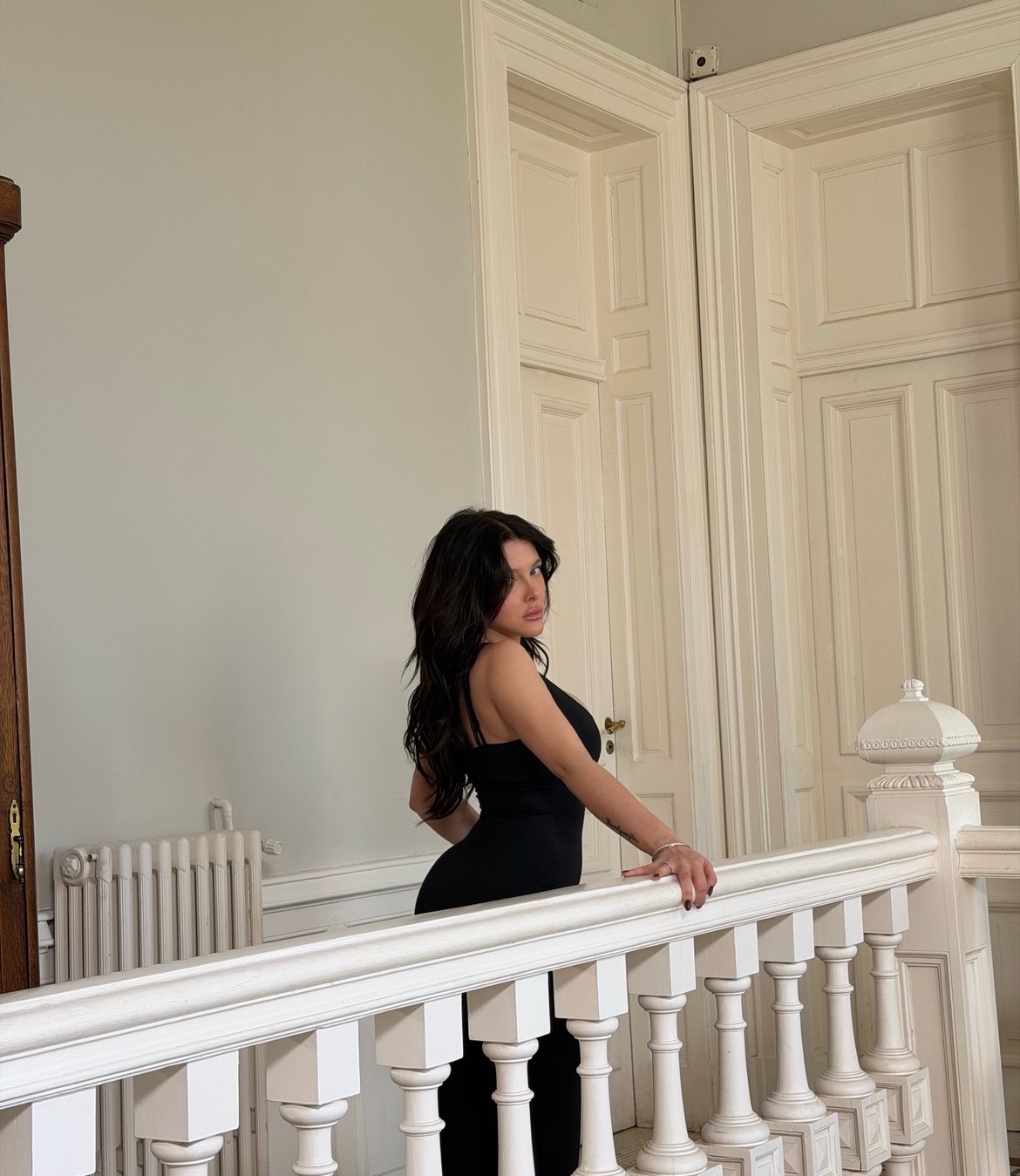Model in a black dress looking back over a white balustrade before tall paneled doors