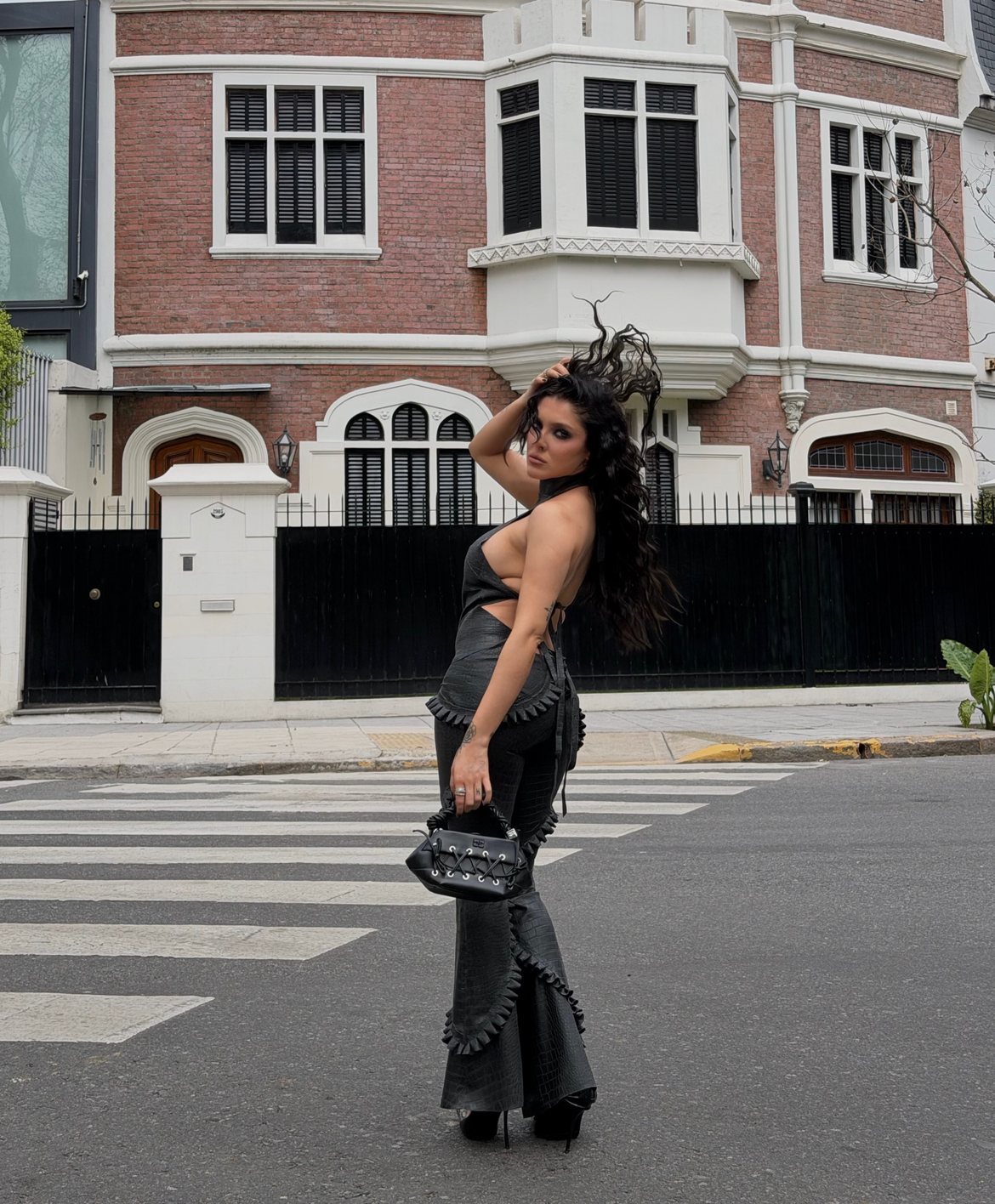 Model in a black croc-leather halter and ruffled flares mid-crosswalk, hair flying, before a red-brick mansion
