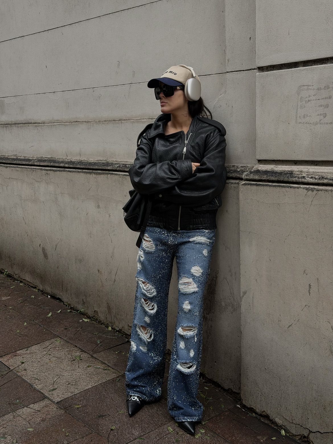 Arms crossed against a stone wall in a leather bomber, Miu Miu cap and crystal-studded jeans