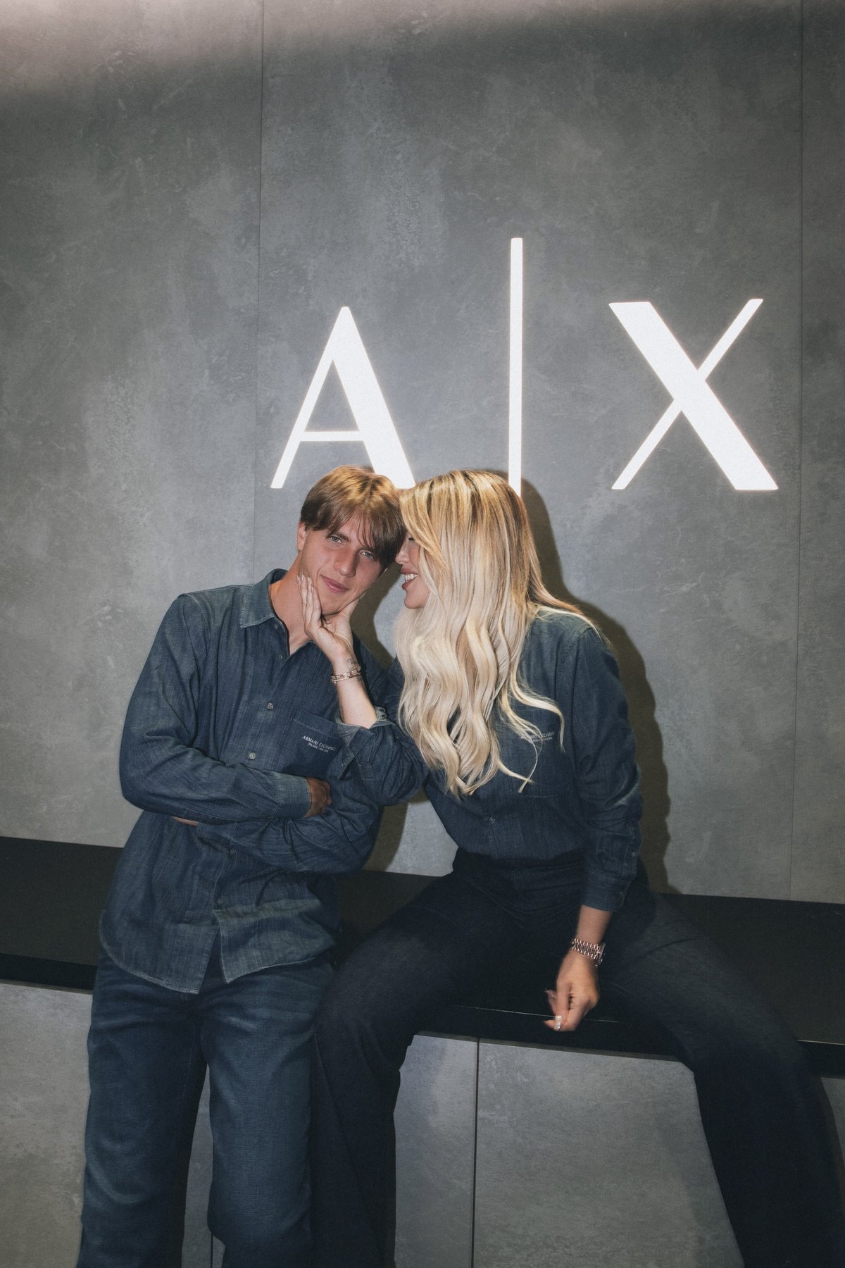 Wanda Nara holding Valentino's face, both laughing, in front of the A|X logo