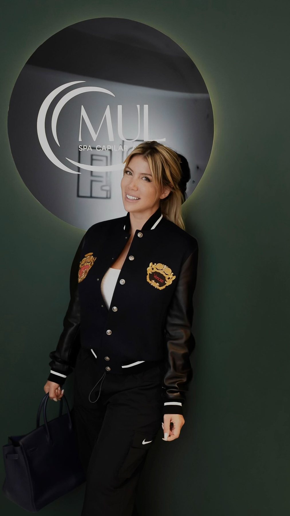 Wanda Nara in a varsity jacket, smiling in front of the backlit MUL spa sign